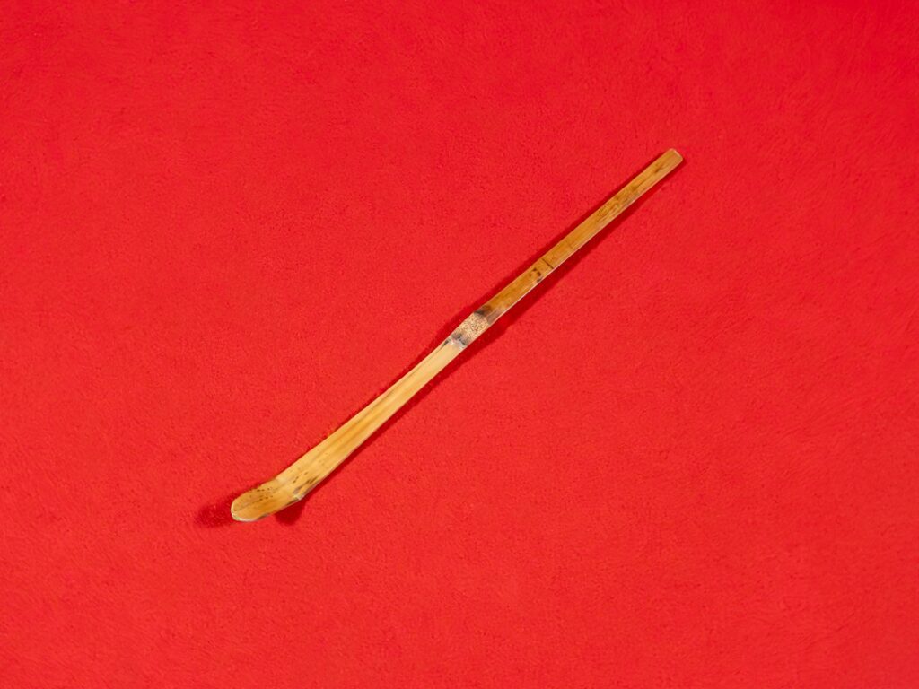 竹製の茶杓が赤い背景に置かれている。査定において重要な素材や状態を視覚的に確認できる構図。共箱や書付の有無も買取価格に影響するため、売却前の確認が重要。